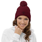 Pom pom beanie in bordeaux met pompon, gedragen door een glimlachende vrouw in een witte hoodie.